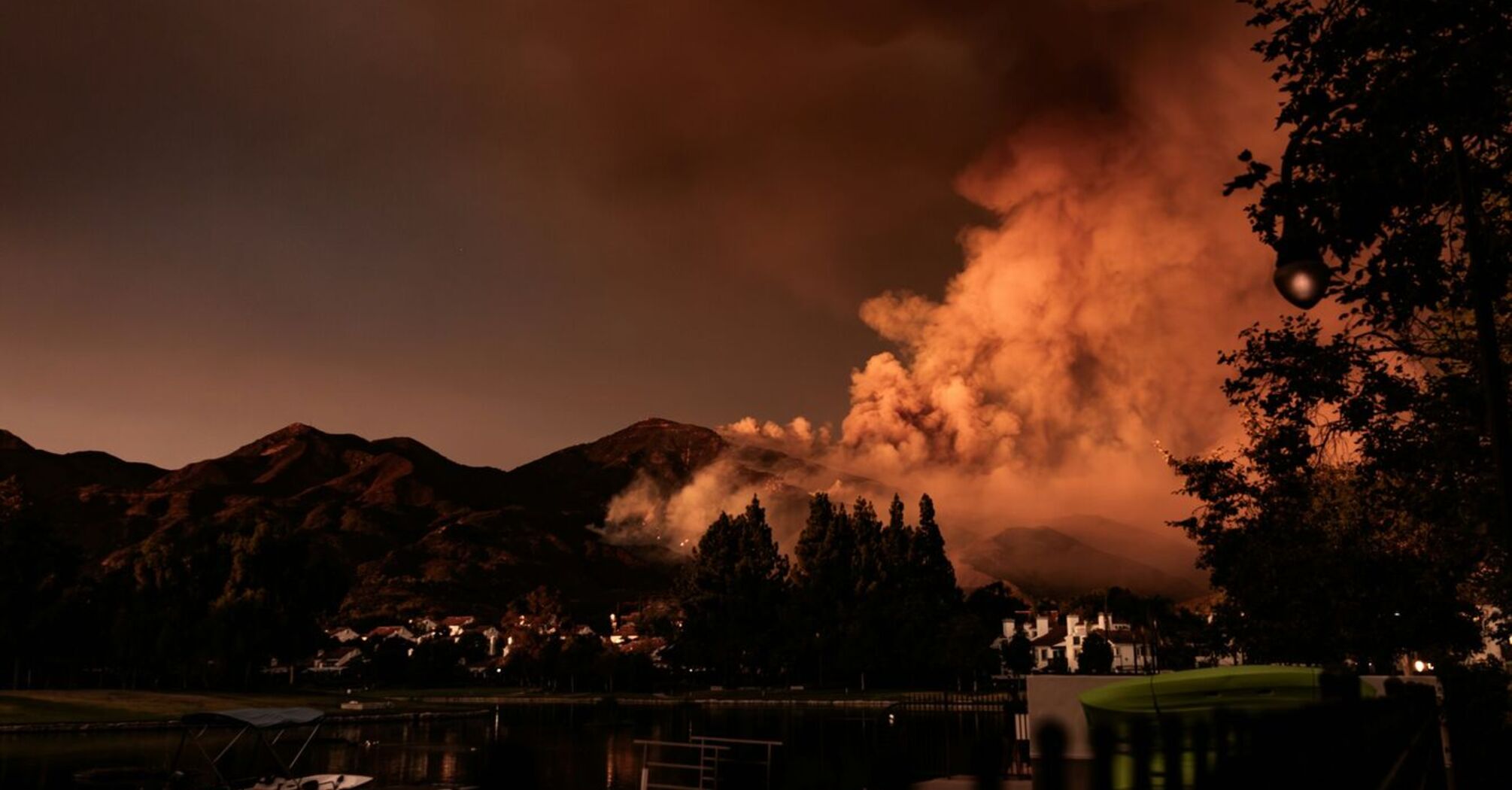 A wildfire burns through a mountainous area at dusk, with heavy smoke rising and a small lakeside community in the foreground