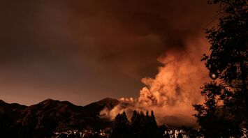 A wildfire burns through a mountainous area at dusk, with heavy smoke rising and a small lakeside community in the foreground