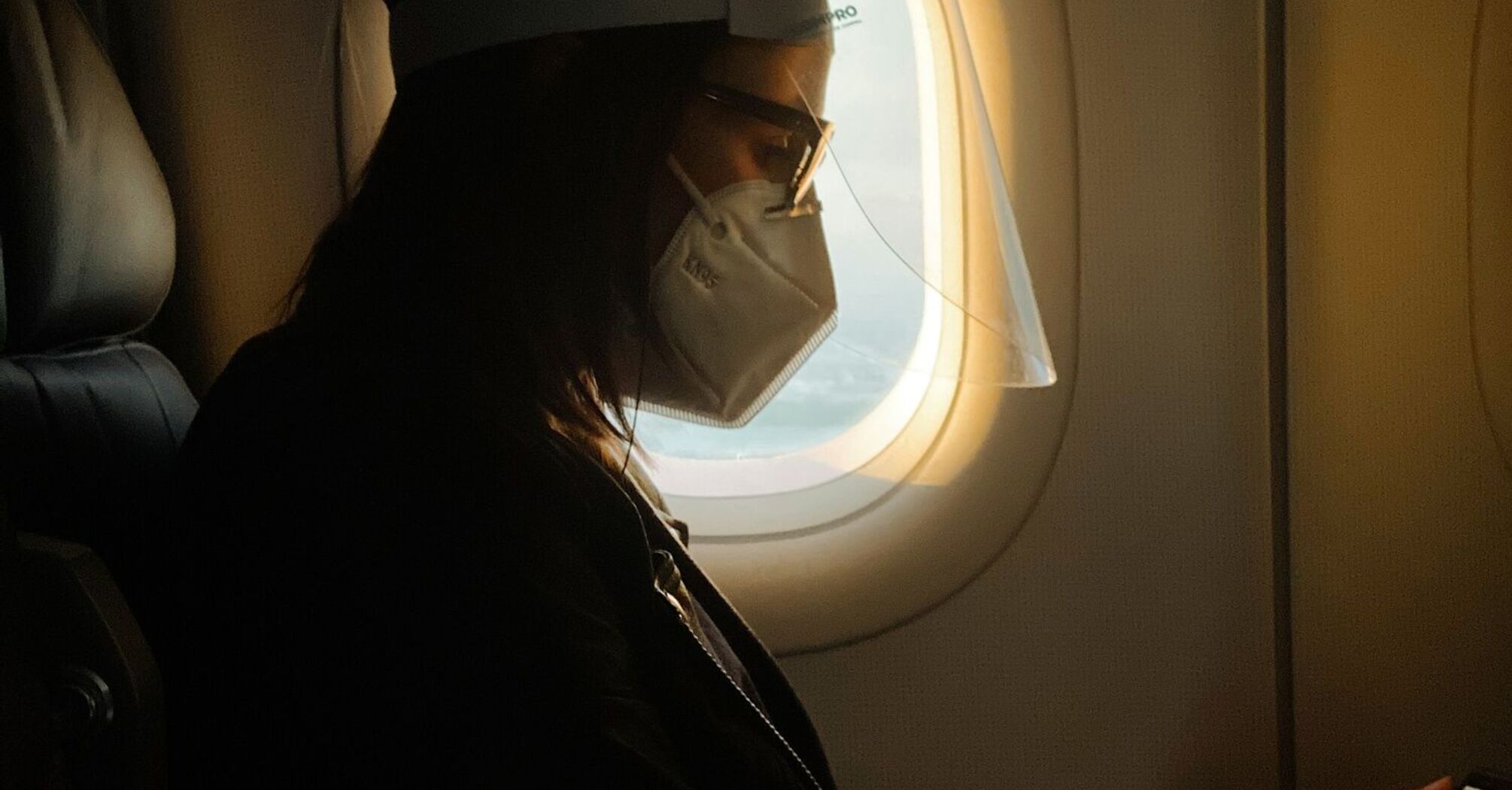 A traveler wearing a mask and face shield seated on a plane while reading on a device