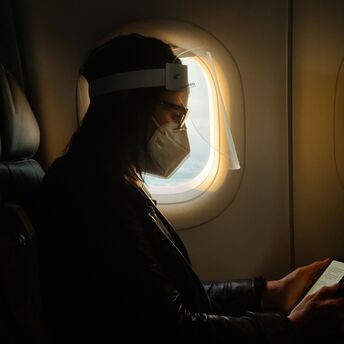 A traveler wearing a mask and face shield seated on a plane while reading on a device