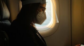 A traveler wearing a mask and face shield seated on a plane while reading on a device