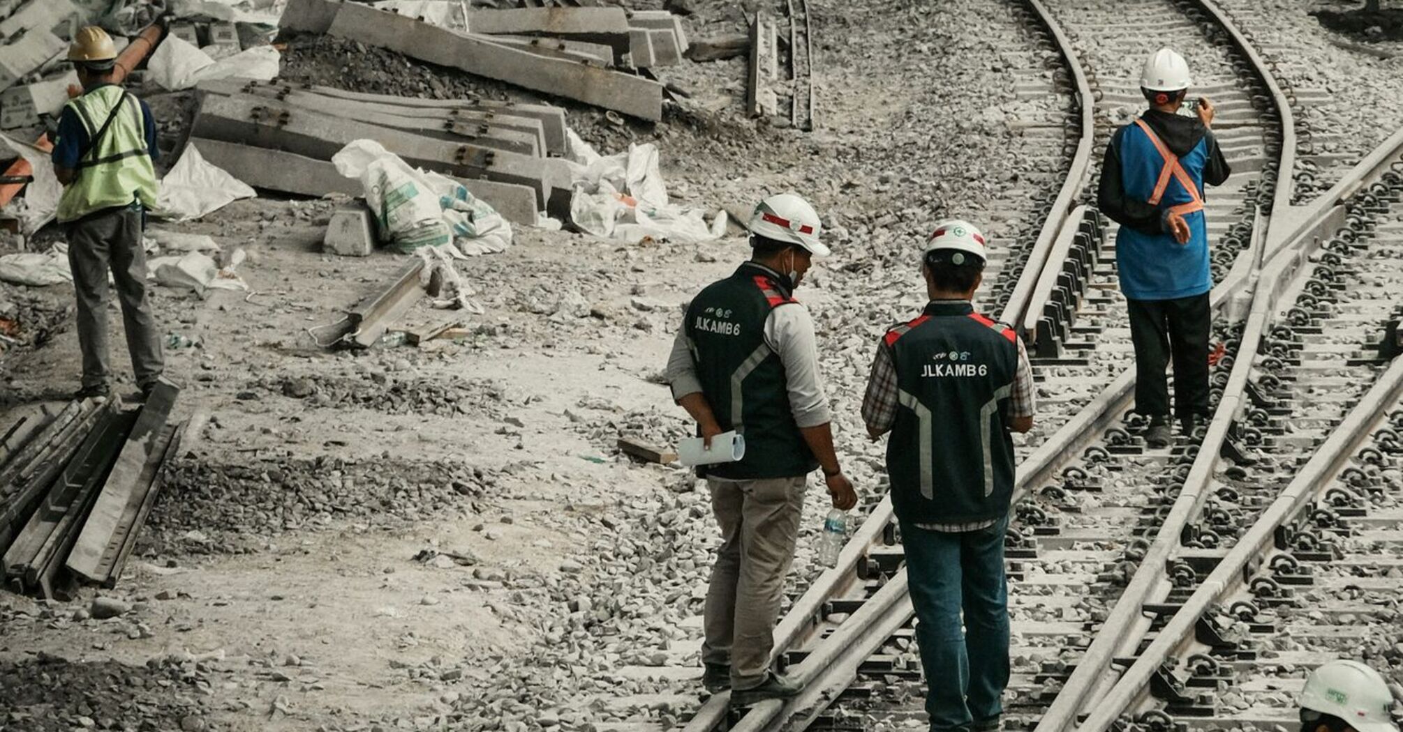 Workers in safety gear conducting maintenance on railway tracks under an urban bridge with construction materials scattered around