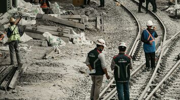 Workers in safety gear conducting maintenance on railway tracks under an urban bridge with construction materials scattered around