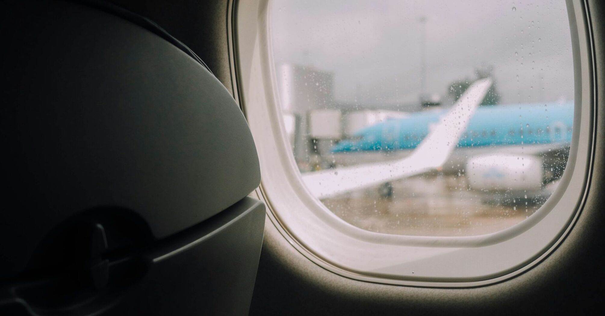 View through an airplane window on a rainy day at the Manchester Airport, with a plane tail visible in the distance