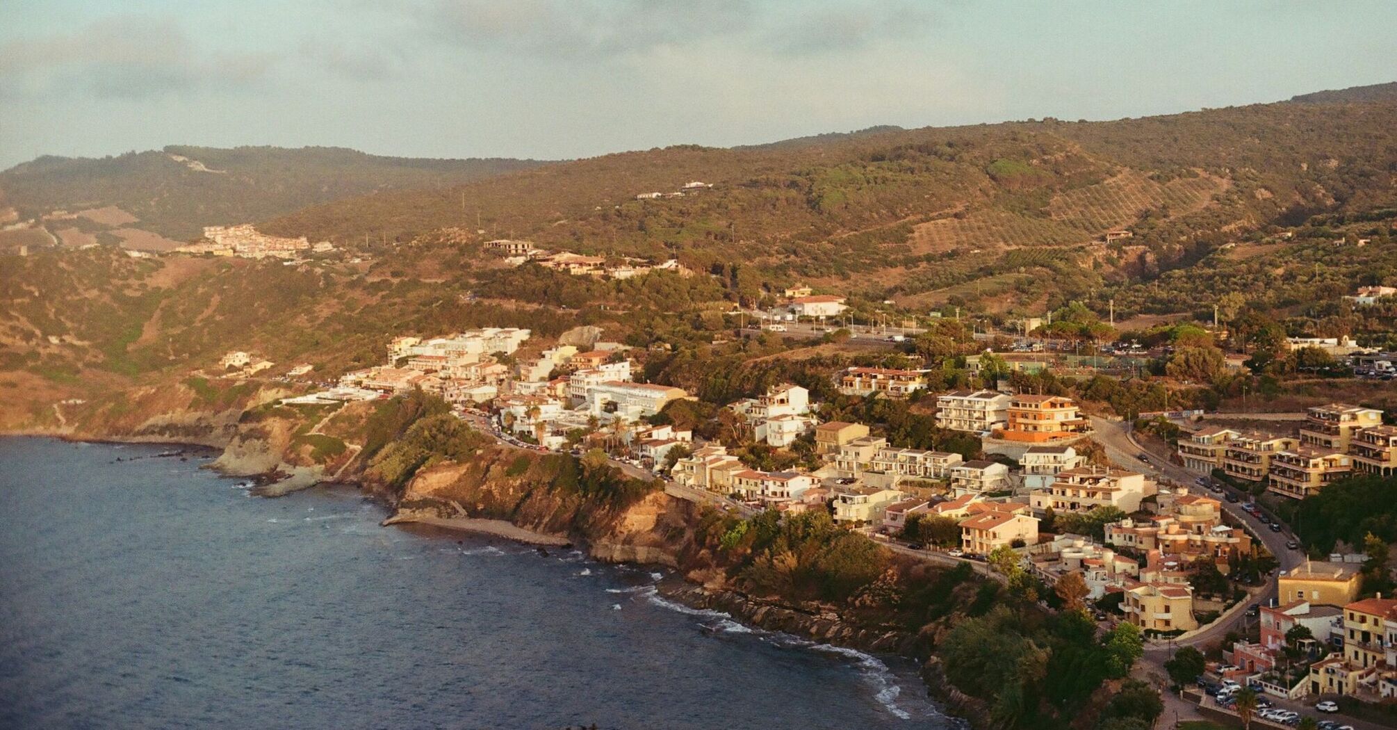 A coastal village in Sardinia with rolling hills and lush greenery, overlooking the Mediterranean Sea under soft evening light