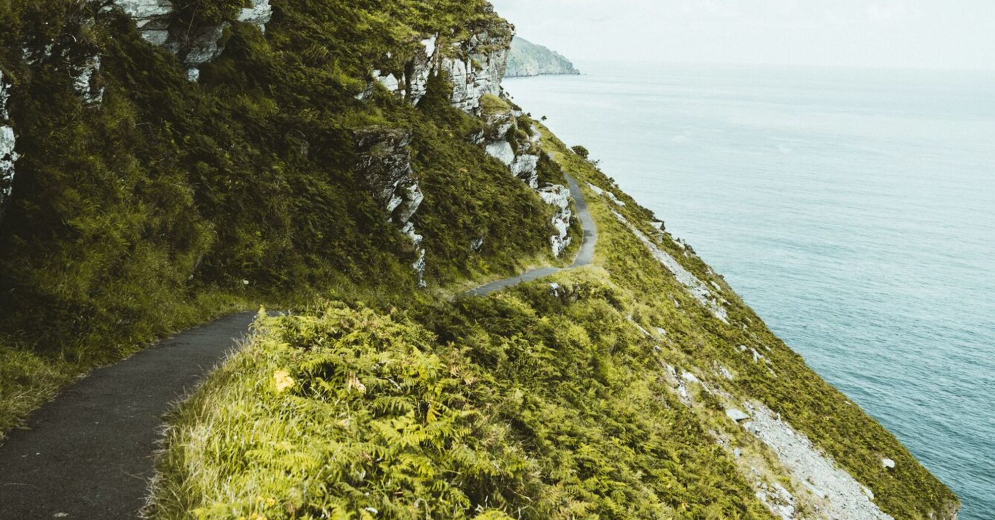 A winding coastal path along green cliffs overlooking the sea under a cloudy sky