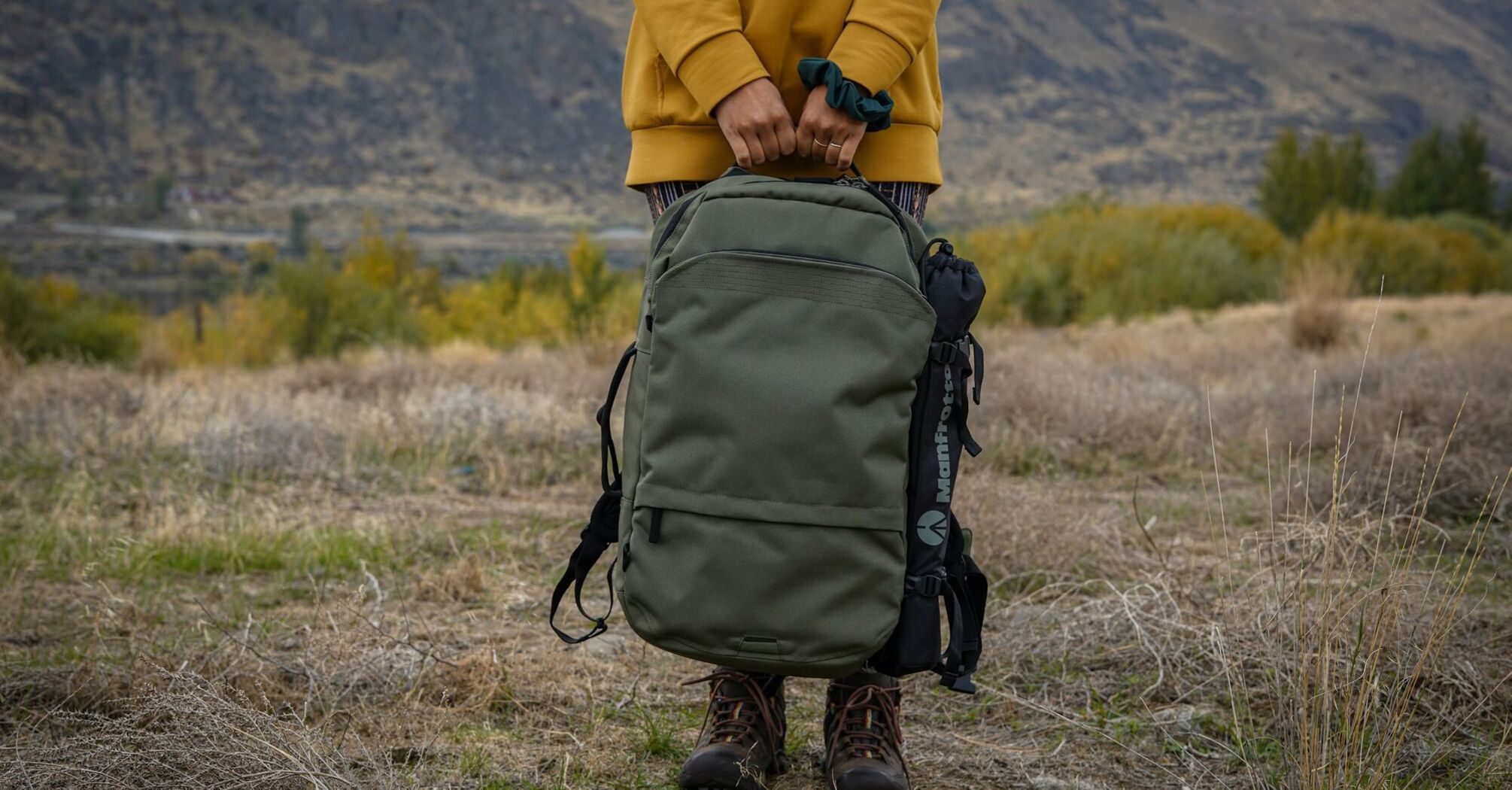 A person holding a green, durable backpack in an outdoor setting, with rugged boots visible on a grassy landscape