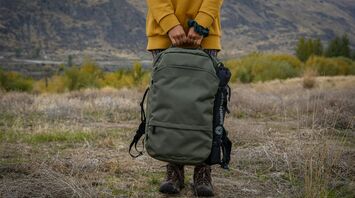 A person holding a green, durable backpack in an outdoor setting, with rugged boots visible on a grassy landscape