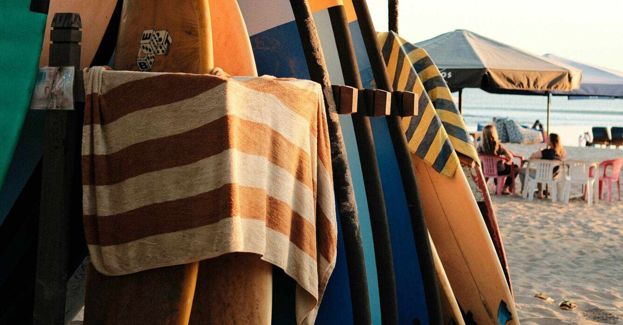 A rack of colorful surfboards on a sandy beach with a striped towel hanging, set under a canopy, against a backdrop of calm ocean and seated beachgoers