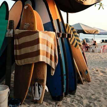 A rack of colorful surfboards on a sandy beach with a striped towel hanging, set under a canopy, against a backdrop of calm ocean and seated beachgoers