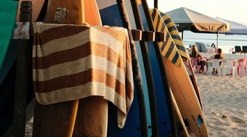 A rack of colorful surfboards on a sandy beach with a striped towel hanging, set under a canopy, against a backdrop of calm ocean and seated beachgoers