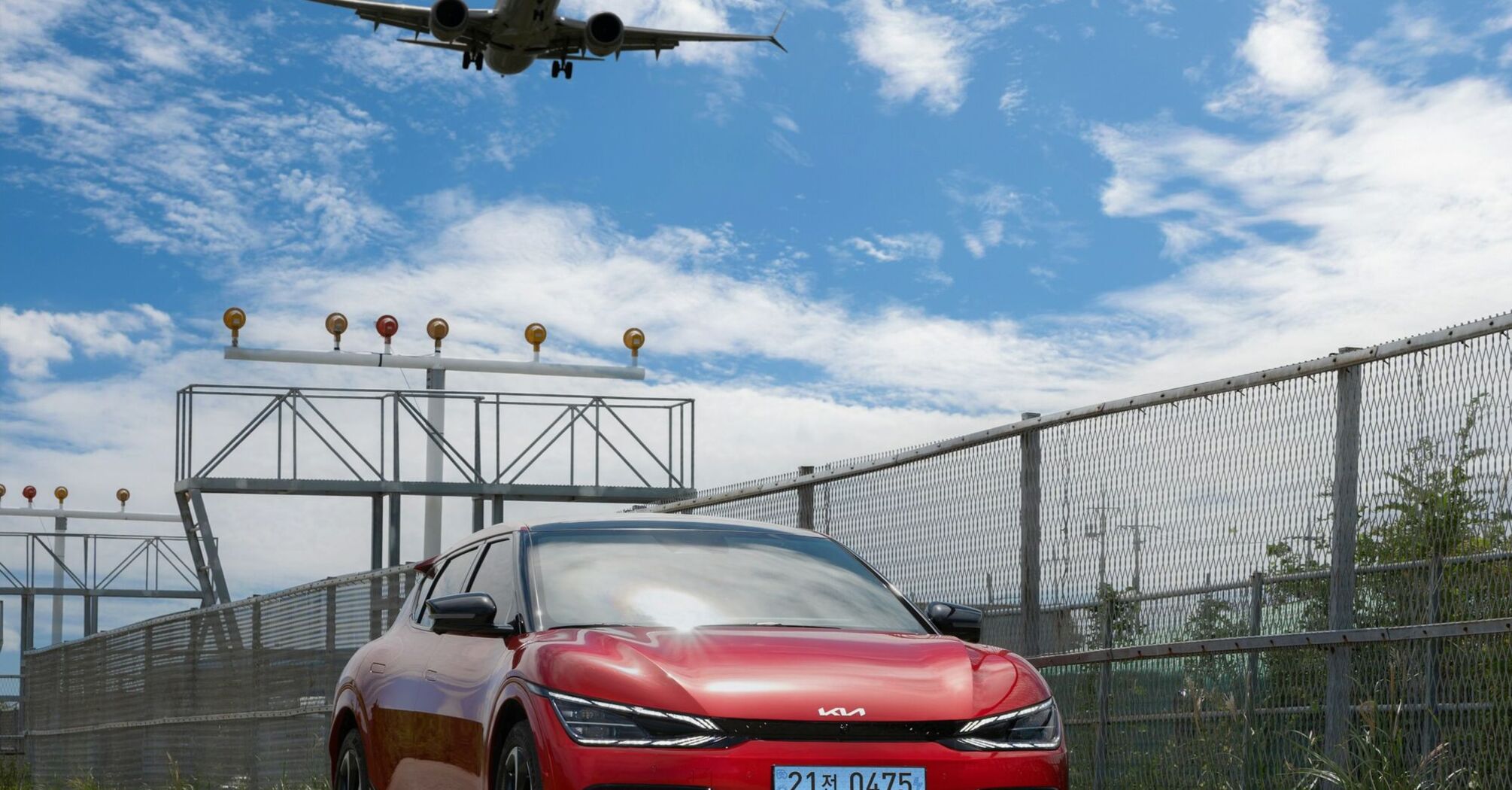 A red car parked near an airport runway with an airplane landing overhead on a sunny day