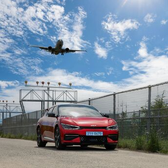 A red car parked near an airport runway with an airplane landing overhead on a sunny day