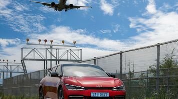 A red car parked near an airport runway with an airplane landing overhead on a sunny day