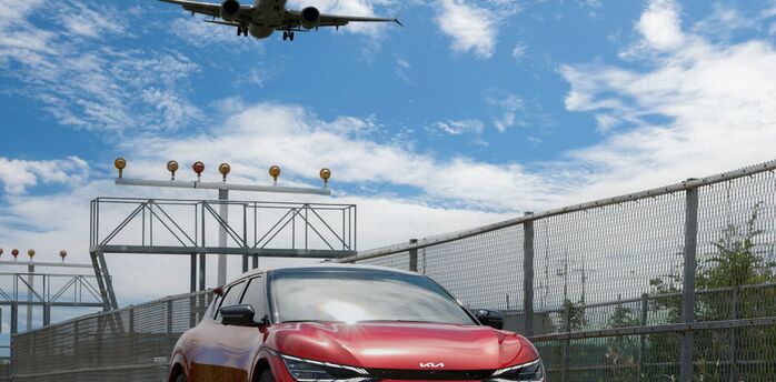 A red car parked near an airport runway with an airplane landing overhead on a sunny day