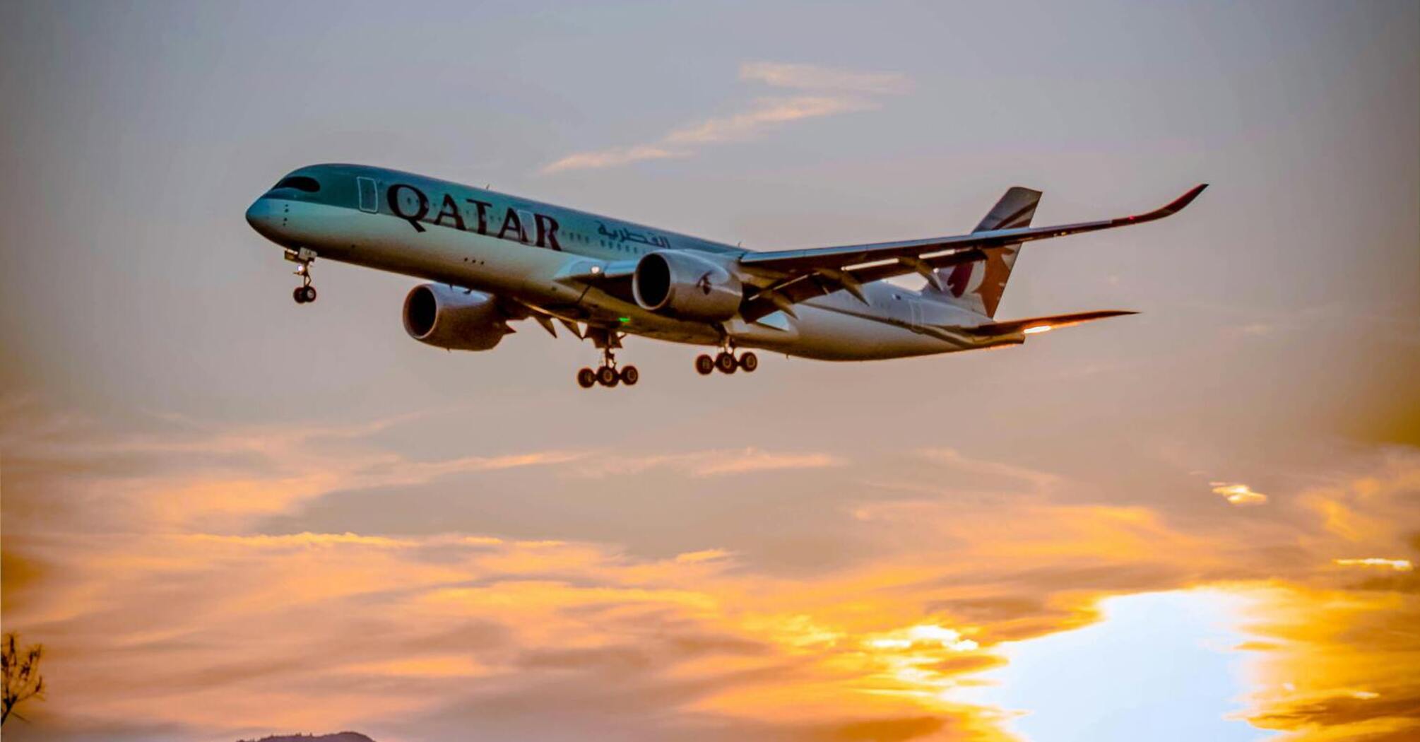 Qatar Airways plane landing against a vibrant sunset with a scenic rural backdrop