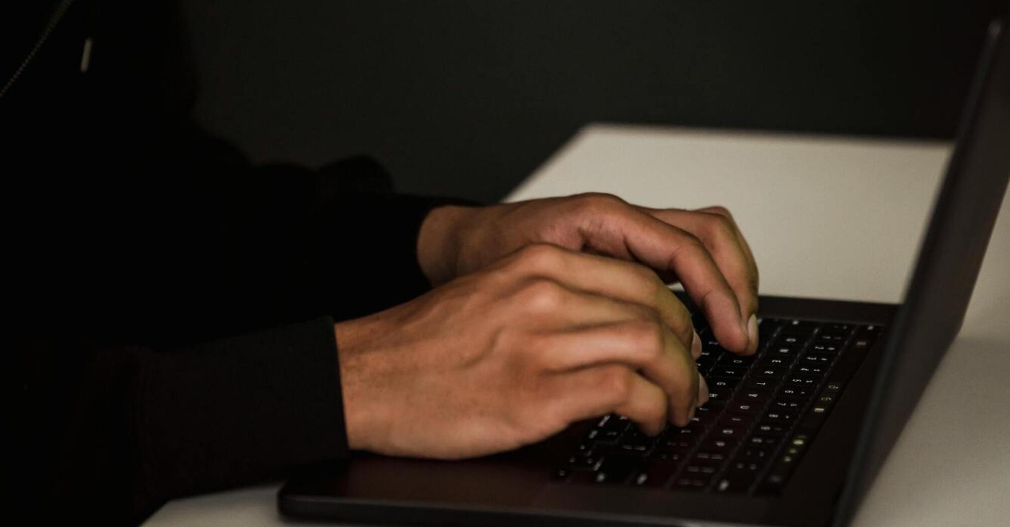 A person in a hoodie typing on a laptop in a dimly lit room