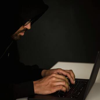 A person in a hoodie typing on a laptop in a dimly lit room