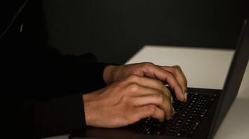 A person in a hoodie typing on a laptop in a dimly lit room