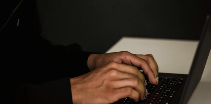A person in a hoodie typing on a laptop in a dimly lit room