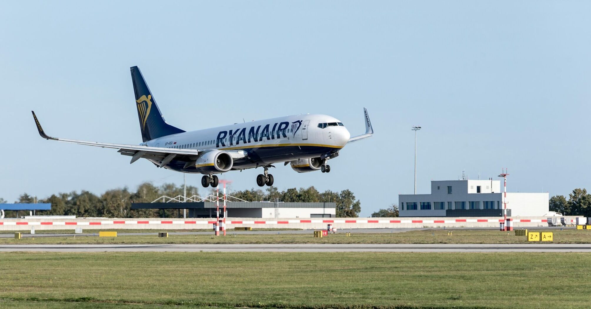 A Ryanair plane landing at an airport, with clear skies and a terminal building in the background