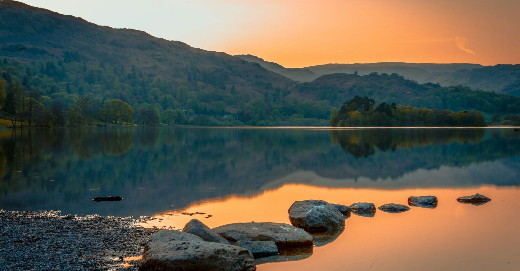 A tranquil lake in the Lake District at sunset, surrounded by rolling hills and reflected in still waters