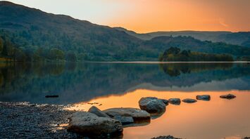 A tranquil lake in the Lake District at sunset, surrounded by rolling hills and reflected in still waters