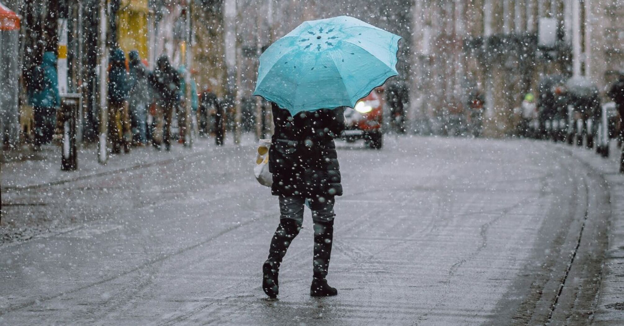 A person holding a turquoise umbrella walks through a snowy street in an urban area