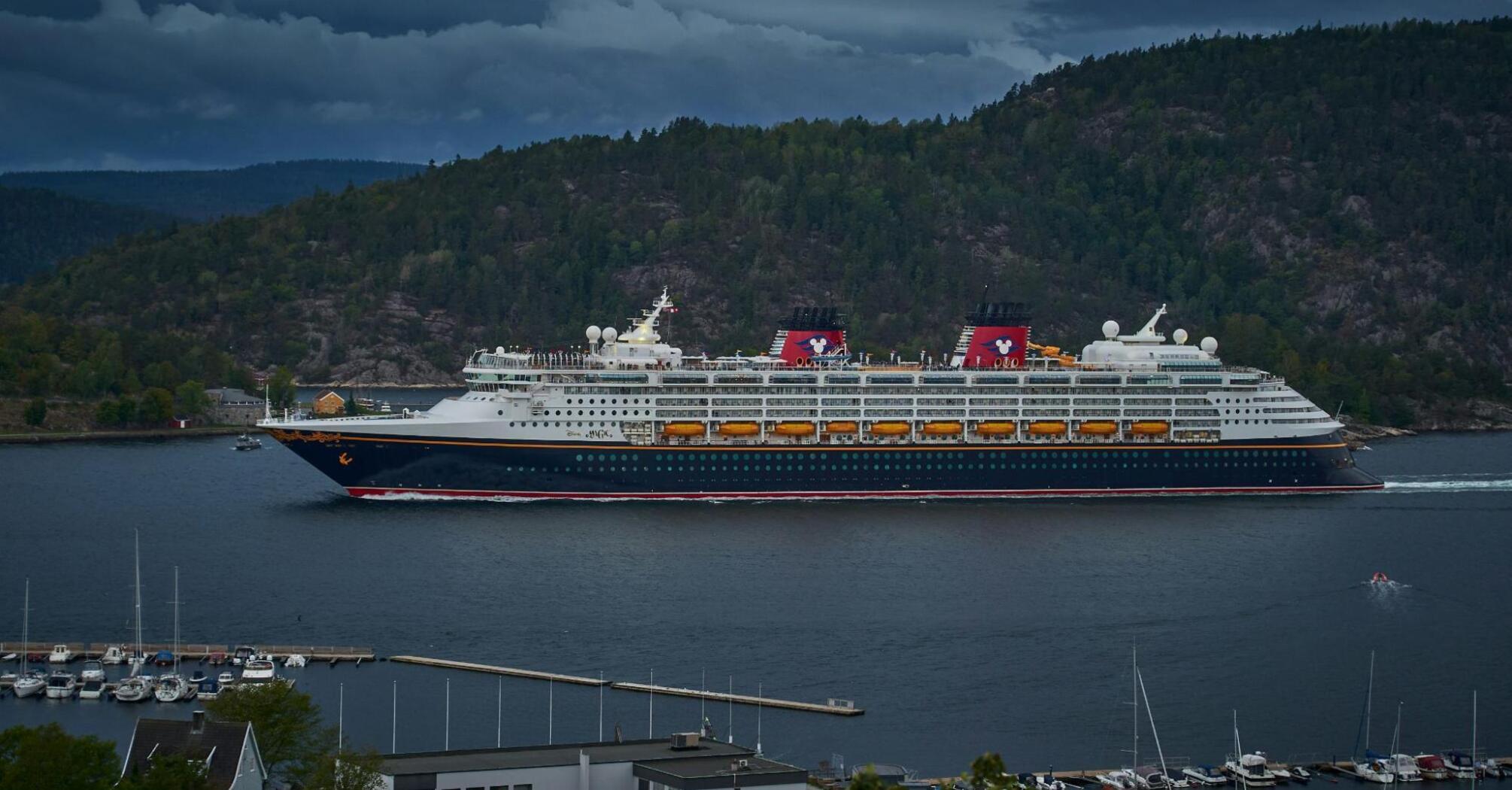 Disney cruise ship sailing through a scenic fjord under a cloudy sky