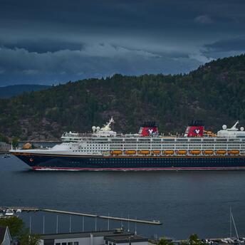 Disney cruise ship sailing through a scenic fjord under a cloudy sky