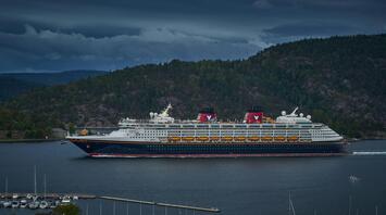 Disney cruise ship sailing through a scenic fjord under a cloudy sky