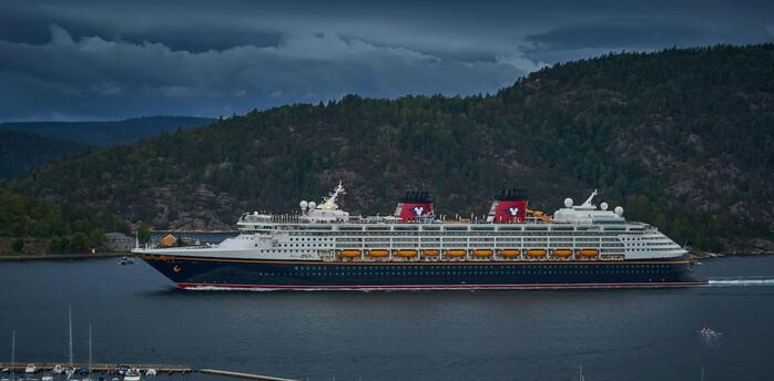 Disney cruise ship sailing through a scenic fjord under a cloudy sky
