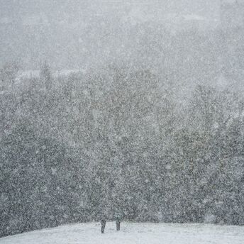 Heavy snowfall obscures a wooded landscape with two people walking through the snow-covered ground