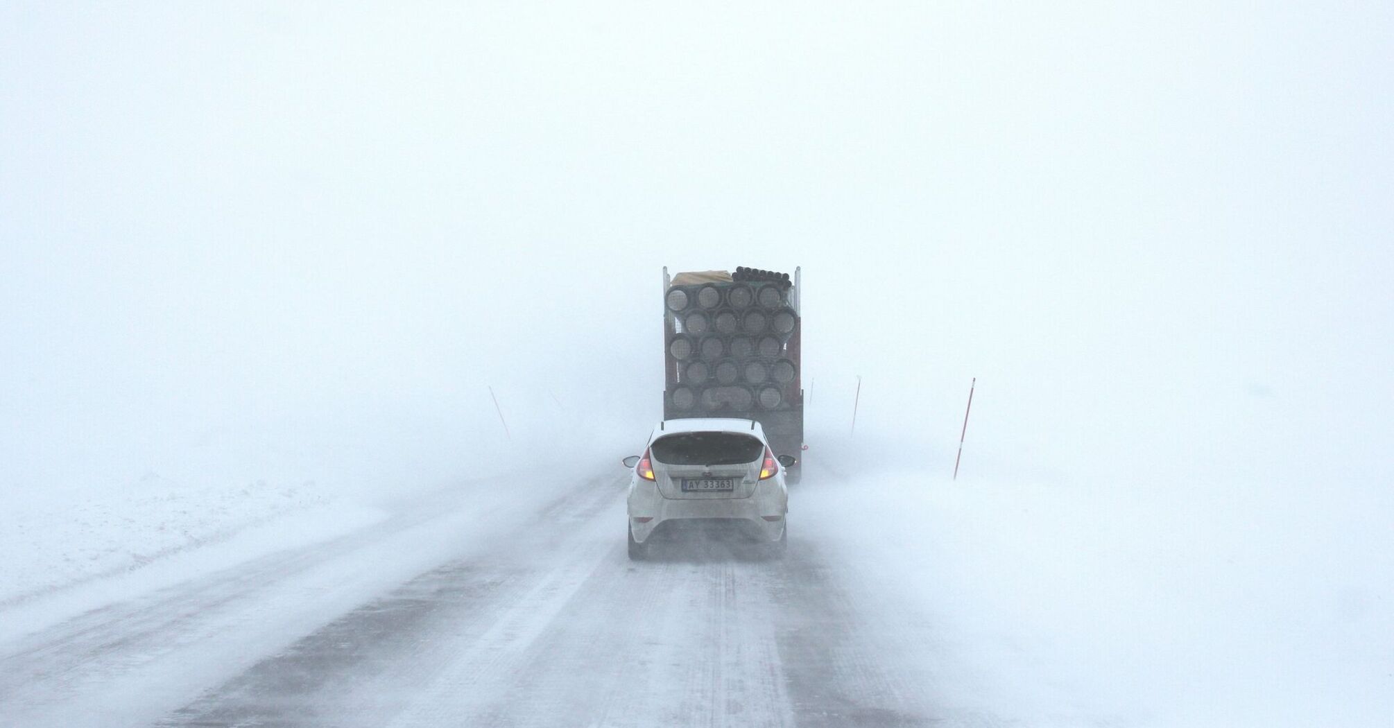 A car and truck driving cautiously on a snowy, fog-covered road with poor visibility