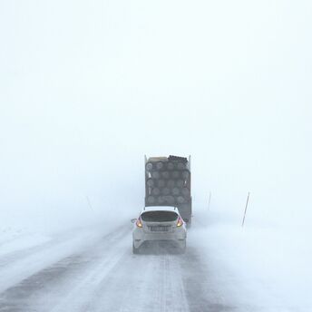 A car and truck driving cautiously on a snowy, fog-covered road with poor visibility