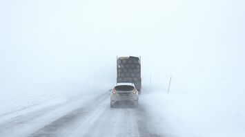 A car and truck driving cautiously on a snowy, fog-covered road with poor visibility