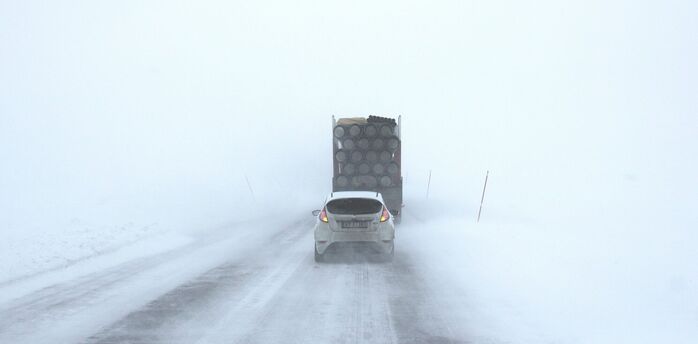 A car and truck driving cautiously on a snowy, fog-covered road with poor visibility