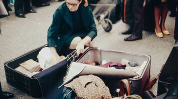 A traveler packing a suitcase outdoors, surrounded by luggage and other travelers