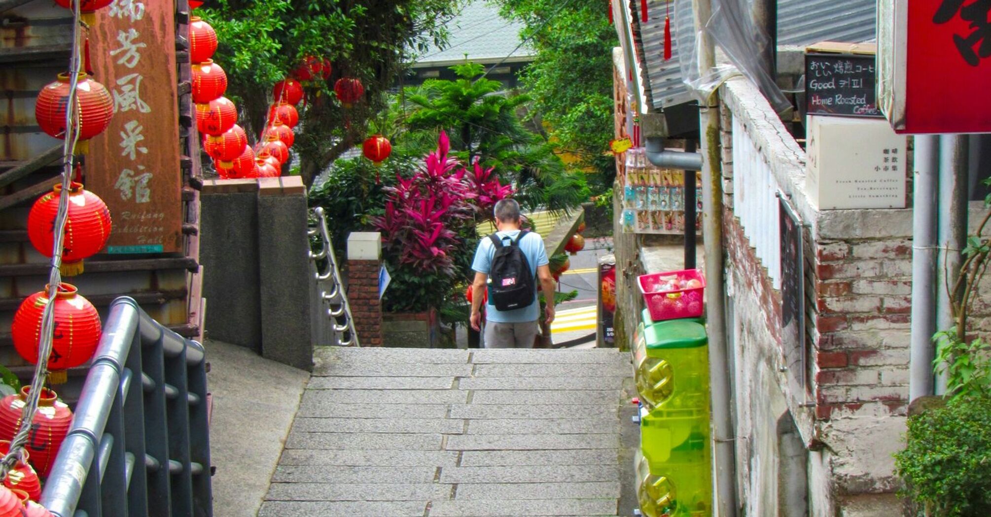 A narrow street in an Asian town adorned with red lanterns, surrounded by greenery and traditional architecture