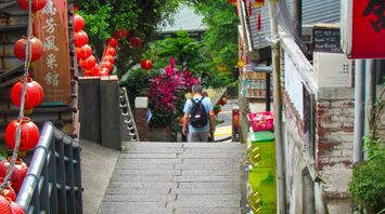 A narrow street in an Asian town adorned with red lanterns, surrounded by greenery and traditional architecture