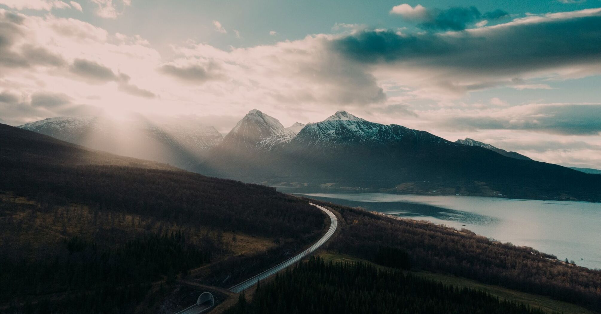 A winding road through a serene mountainous landscape with a glowing sun breaking through clouds, overlooking a calm lake and dense forest