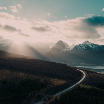 A winding road through a serene mountainous landscape with a glowing sun breaking through clouds, overlooking a calm lake and dense forest