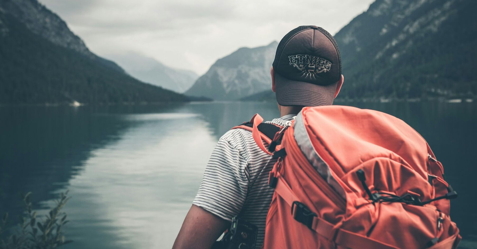 A traveler with an orange backpack overlooks a calm lake surrounded by mountains under an overcast sky