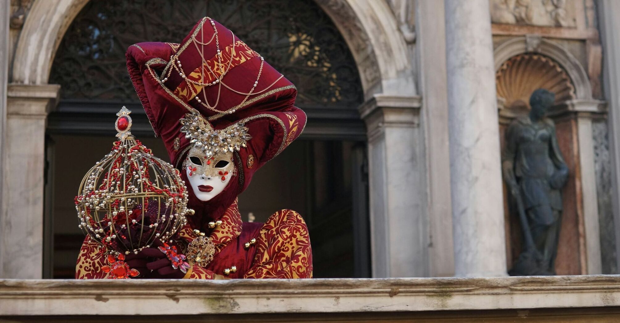 A person in an elaborate Venetian carnival mask and costume holding a decorative object on a marble balcony