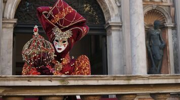 A person in an elaborate Venetian carnival mask and costume holding a decorative object on a marble balcony