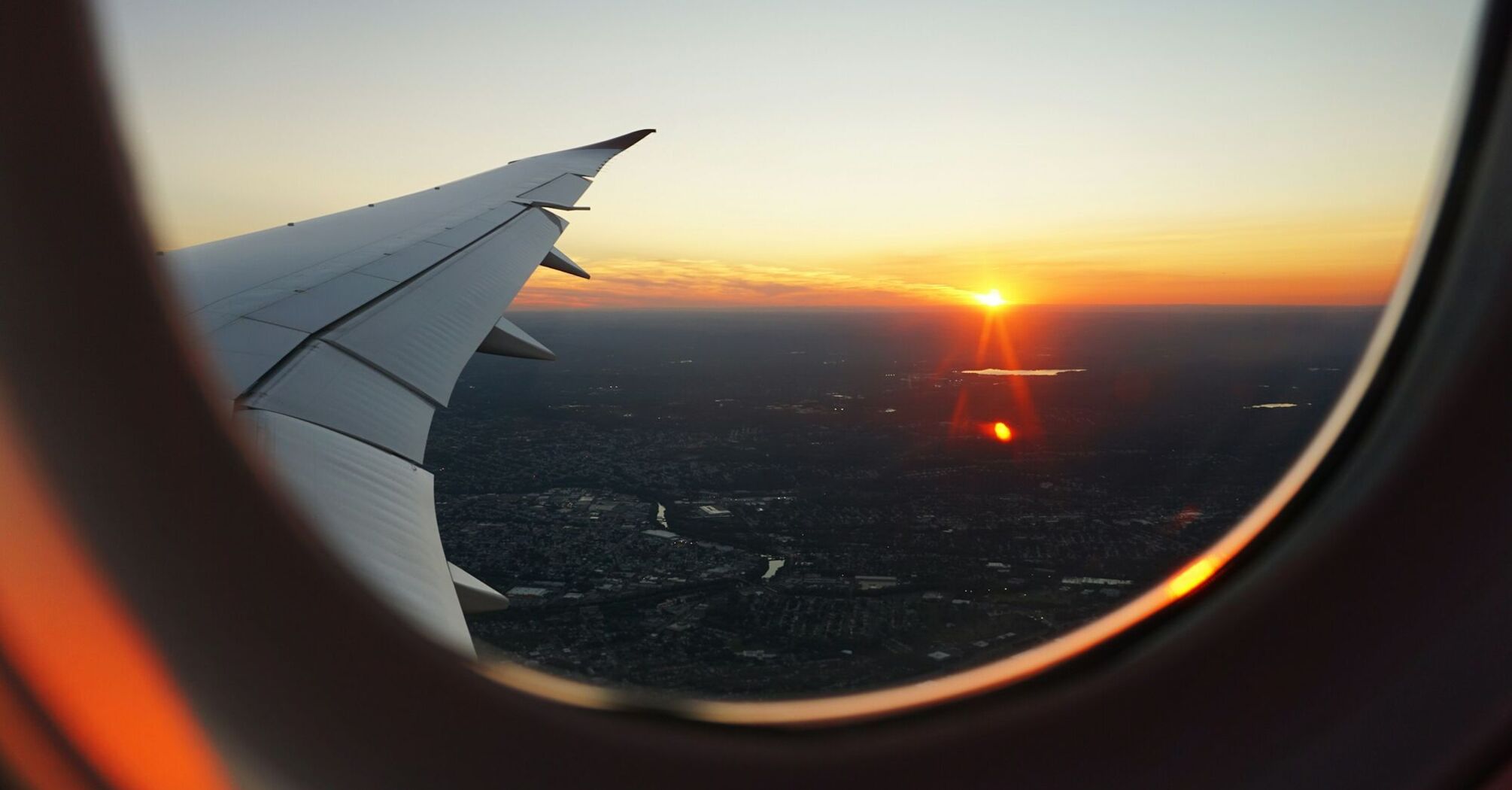 A view of a sunset from an airplane window, showing the aircraft's wing and a city below