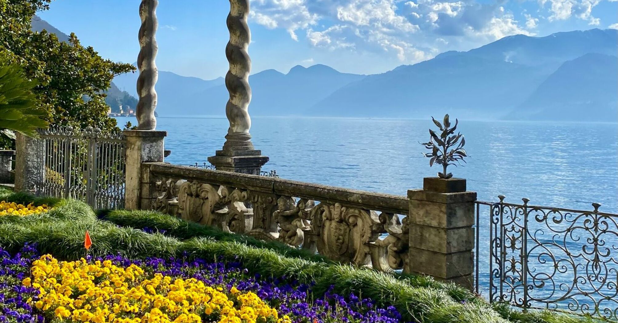 A picturesque lakeside garden with blooming flowers, ornate stone columns, and a view of misty mountains under a clear blue sky