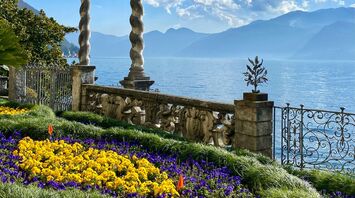 A picturesque lakeside garden with blooming flowers, ornate stone columns, and a view of misty mountains under a clear blue sky