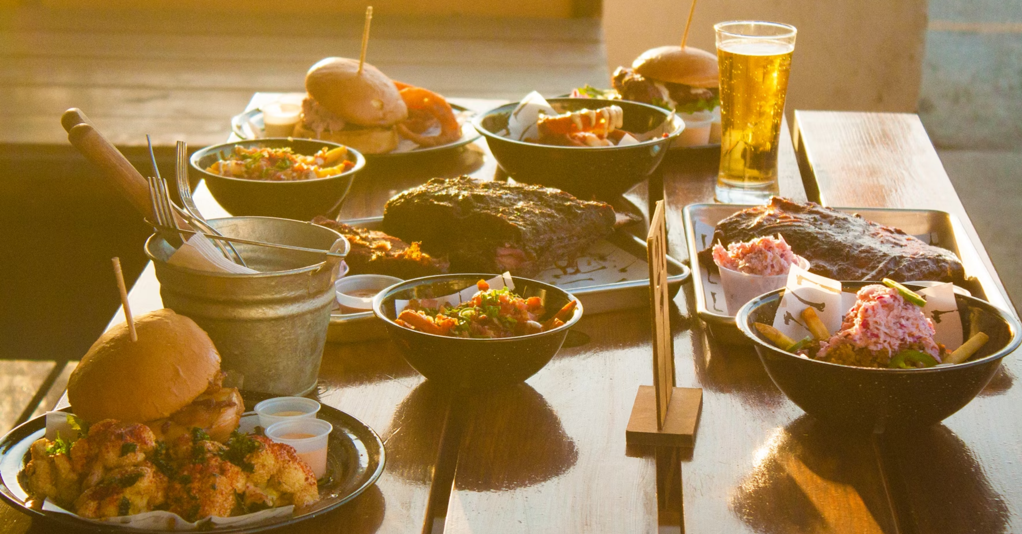 A wooden table with various dishes, including burgers, ribs, coleslaw, and a glass of beer, served in a sunlit outdoor setting