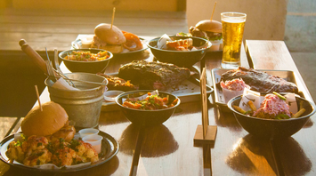 A wooden table with various dishes, including burgers, ribs, coleslaw, and a glass of beer, served in a sunlit outdoor setting